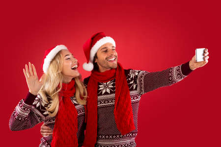 Christmas fun. Romantic young couple in Santa hats taking selfie over red background, panorama