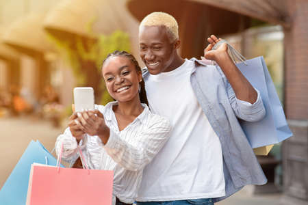 Walking together, shopping and selfies in city. Cheerful african american guy and woman in casual, with paper bags, make photo on smartphone, outdoorsの写真素材