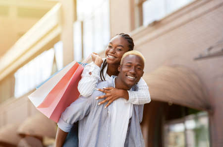Leisure, free time and purchase. Smiling african american couple in casual with paper bags having fun near store, sun flareの写真素材