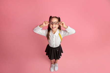 Success in school. Smiling little schoolgirl with glasses and backpack shows tongue and have fun, isolated on pink background, studio shootの写真素材