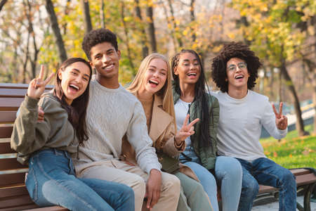 Positive international teenagers enjoying weekend at public park, sitting on bench and posing at cameraの写真素材