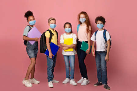 Education during virus outbreak. Diverse children with schoolbags and notebooks wearing protective masks on pink backgroundの写真素材