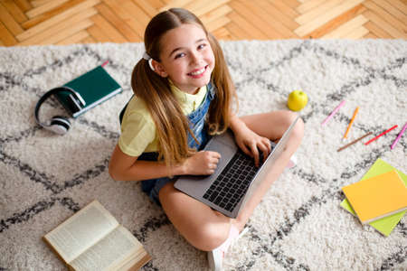 Above view portrait of smiling little teenage girl sitting on the floor carpet, using laptop and looking up at cameraの写真素材