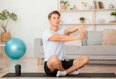 Sports lifestyle at home. Attractive man doing stretching for hand exercises on mat in living room...