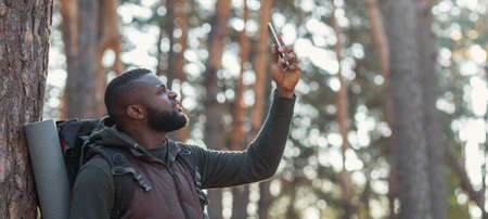 African young man hiker looking for mobile network in forest, holding smartphone in his hand, panorama with copy spaceの写真素材