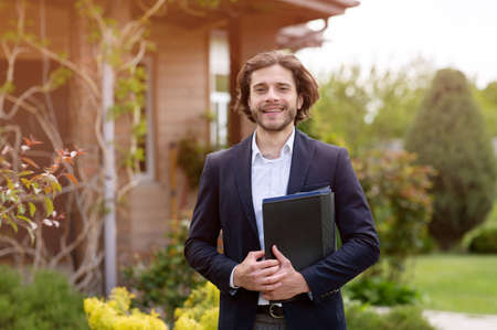 Portrait of happy real estate agent with documents posing near beautiful house outdoorsの写真素材