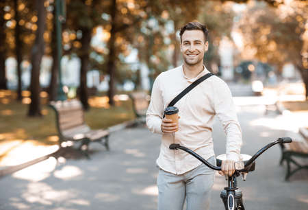 Way to office. Smiling guy in formal wear with cup of coffee walking with bike in park, copy spaceの写真素材