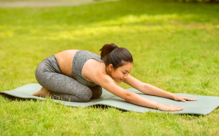 Calm Asian woman doing child yoga pose on sports mat at green park, panorama