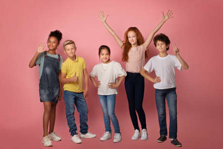 Full length portrait of happy diverse schoolkids making different gestures over pink background