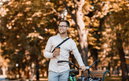Modern lifestyle and ecological transport. Happy hipster in glasses holding coffee and walking with bike in park, copy spaceの写真素材