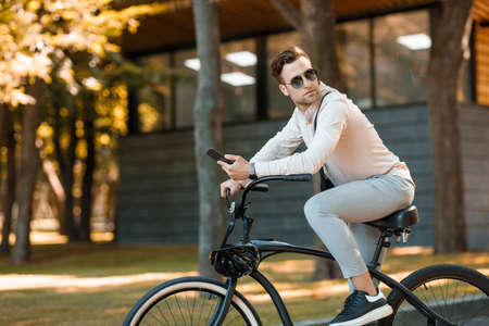 Businessman riding on bicycle to work in morning. Guy in sunglasses and smartphone in the park, copy spaceの写真素材