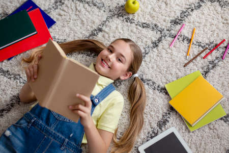 Leisure Concept. Top view of cute smiling girl lying on floor, holding paper book, looking up at...