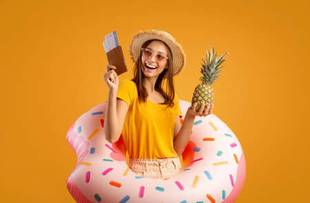 Cheerful girl with sunglasses and summer hat holding pineapple and passport, wearing rubber ring, yellow studio backgroundの写真素材