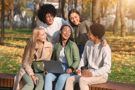 Cheerful multiethnic friends having fun at public park, looking at laptop screen and laughing
