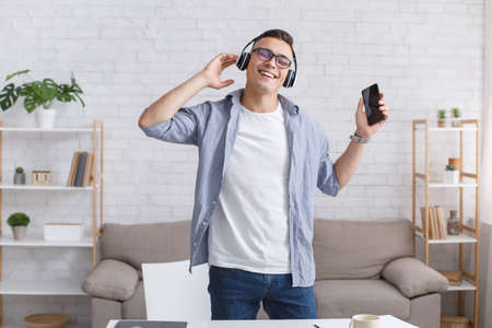 Favorite music during break at remote work at home. Happy guy in headphones holds smartphone and...