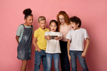 Surprised schoolkids watching shocking news on smartphone against pink backgroundの写真素材