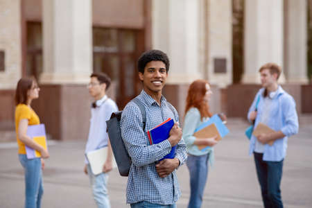 Happy African American Student Guy Posing With Books And Backpack Standing Near University Building Outdoor. Study Abroad Conceptの写真素材