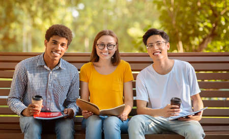Three Multiethnic College Students Sitting On Bench Relaxing After Classes, Learning And Having Coffee Outdoor, Smiling To Camera.の写真素材