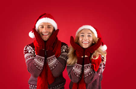 Its Christmas time. Adorable young couple in mittens, Santa hats and scarves on red backgroundの写真素材