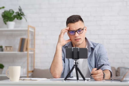Webinar and online meeting during quarantine. Pensive guy with glasses makes notes and watches in webcam on smartphone in interior of home, copy spaceの写真素材