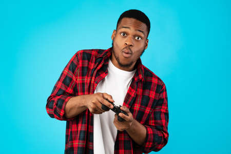Black Millennial Guy Playing Video Games Holding Gamepad Standing Over Blue Studio Background....