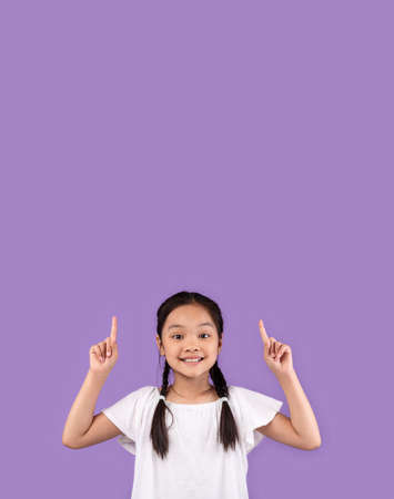Look Up. Adorable Asian Kid Girl Pointing Fingers Upward At Copy Space Standing Over Purple Studio...