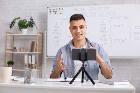 Teacher recording vlog. Attractive young man explains english rules and gestures on webcam in class at homeの写真素材
