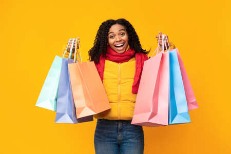 Autumn Sales. Excited Black Girl With Shopping Bags Posing Wearing Scarf And Winter Jacket Over Yellow Studio Backgroundの写真素材