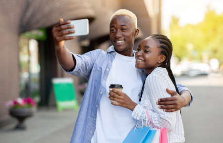 Selfie for a walk. Happy african american guy makes photo and hugs woman, girl holds takeaway coffee, outdoors, copy spaceの写真素材