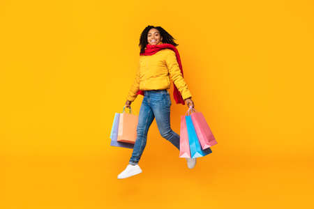 Winter Shopping And Sales. African Girl With Shopping Bags Jumping In Mid-Air Posing Wearing Warm...