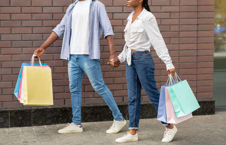 Shopping in mall on weekends. African american couple with multicolored bags go away from store,...
