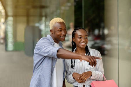 Cool, excellent discounts. Happy african american guy pointing on showcase of mall, to girl with colored bags, outdoors, free spaceの写真素材