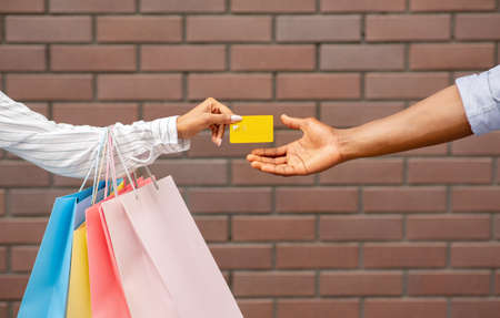 Shopping in boutique. African american girl with multicolored packages gives credit card to seller on brick wall background, cropped, free spaceの写真素材