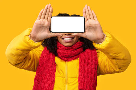 African American Woman Holding Phone With Empty Screen In Front Of Eyes Standing On Yellow Studio...