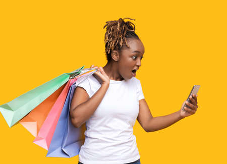 Amazed african american girl with colorful shopping bags looking at mobile phone screen, finally...