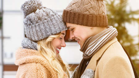 Portrait of romantic young couple in knitted hats touching foreheads during winter date outdoors,...