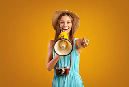 Female tourist with megaphone pointing at camera, yellow studio background, excited deals for...