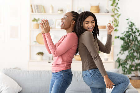 Cheerful black girlfriends singing and dancing, having fun together at homeの写真素材