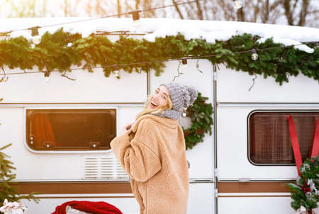 Winter Joy. Beautiful blonde girl posing near rv at campsite in wood, having fun, laughing at camera, enjoying christmas holiday season, wearing coat and woolen hat, feeling happy, copy spaceの写真素材