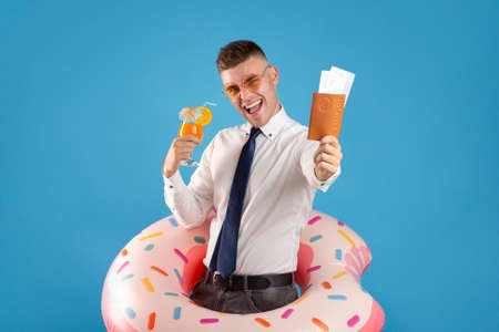 Excited young businessman with passport, plane tickets, inflatable ring and refreshing cocktail ready for summer vacation, blue backgroundの写真素材