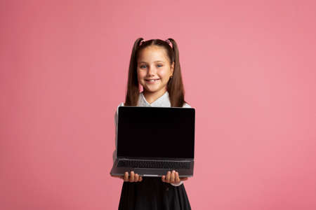 Remote education during quarantine. Smiling girl in school uniform holding laptop with blank screen, isolated on pink backgroundの写真素材