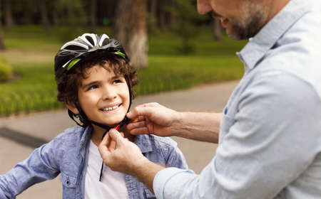 Safety First. Closeup of father putting protection helmet on his little son at park, outdoor shot