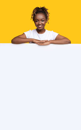 Cheerful black woman leaning on empty board for arvertisement, yellow backgroundの写真素材