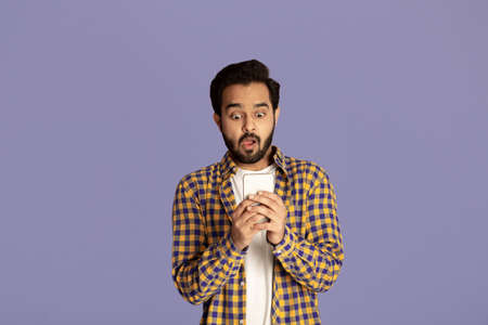 Handsome Indian man looking at his smartphone in shock over lilac backgroundの写真素材