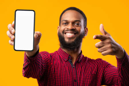 Trendy Mobile Phone. Smiling bearded black man holding latest slim smartphone with white blank screen in hand and pointing at it, indicating empty space for mock up, blurred backgroundの写真素材