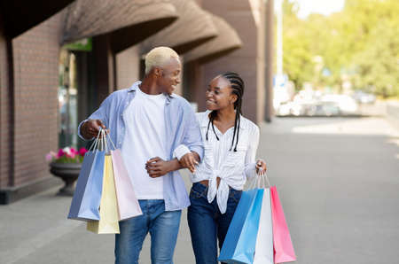 Travel and shopping in city. Cheerful african american young man and woman with colored bags go from mall and discuss purchasesの写真素材