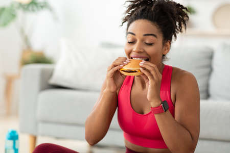 Cheat Meal. Black Fit Woman Eating Burger Enjoying Unhealthy Fast Food After Training Sitting On...