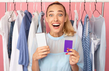 Excited overjoyed woman holding smartphone and purple credit card in hands, ordering stylish clothes and purchases online via internet, standing with rack in the pink backgroundの写真素材