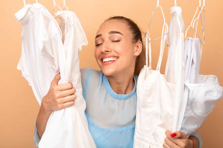 Portrait of happy woman choosing new outfit, standing in rack with hangers, hugging clothes. Satisfied consumer in love with shopping, excited about apparel, beige backgroundの写真素材