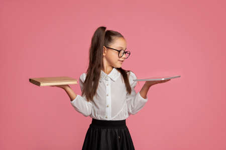 Books or modern technology. Elementary school girl comparing tablet and book on pink background, studio shotの写真素材